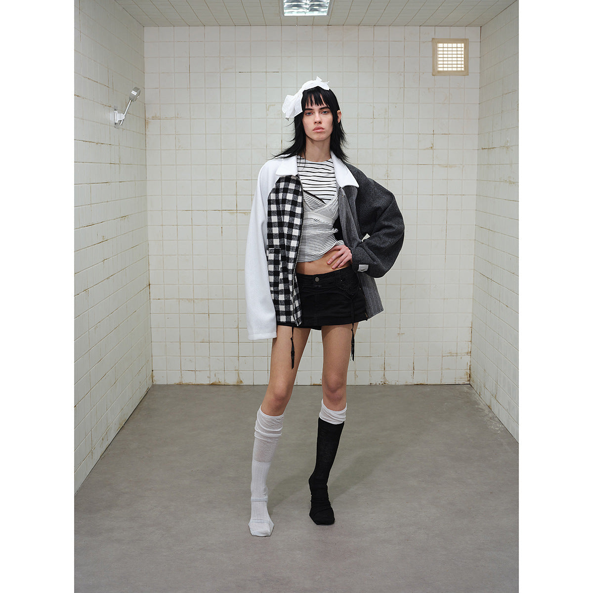 Full-length model wearing a patchwork jacket with white sleeves, check and gray pinstripe panels, posed in a tiled room.