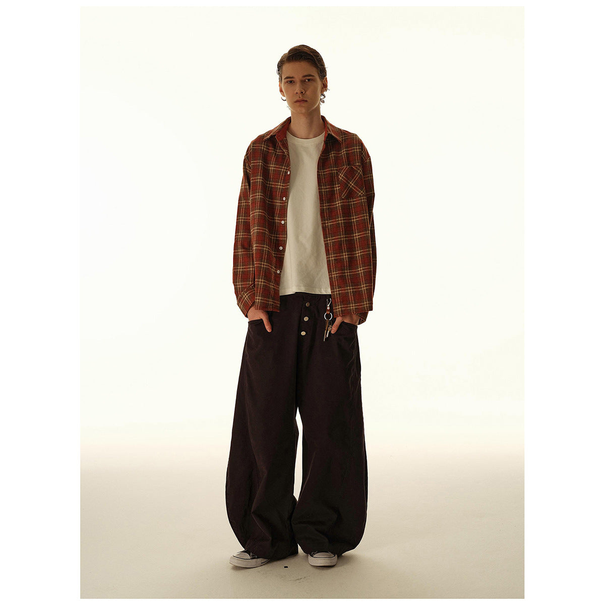 Full-length model wearing an open rust plaid shirt over a cream tee, with very wide black trousers.