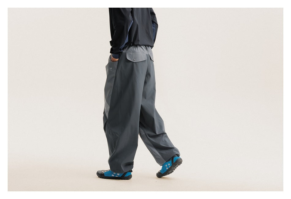 Rear three-quarter view of a model wearing loose charcoal-gray pants with a button-flap back pocket and contrast panels.