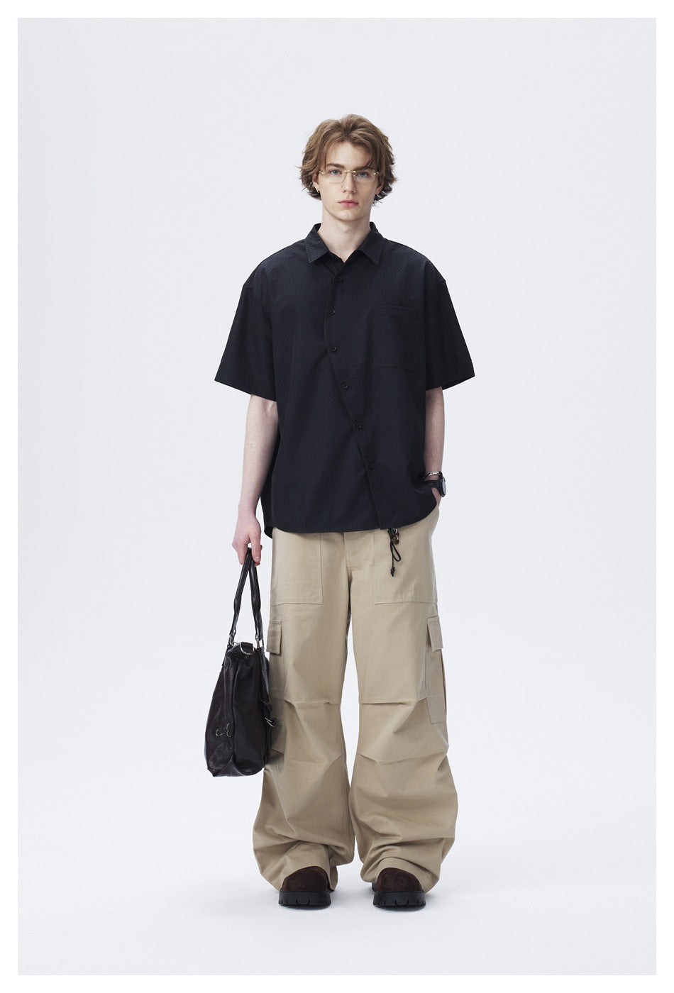 Model wearing a black short-sleeve shirt with a diagonal button front over wide khaki cargo pants, holding a bag