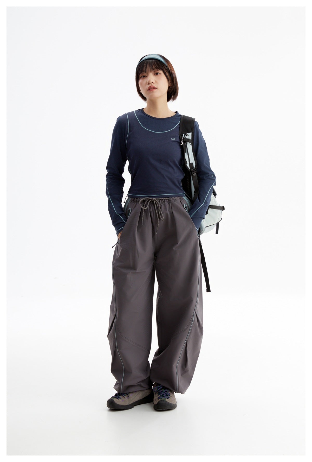 Model wearing a navy long-sleeve top with turquoise piping and charcoal wide-leg pants, carrying a backpack.