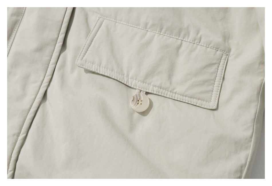 Close-up of an ivory coat’s flap pocket and button closure with the garment’s stitching visible.