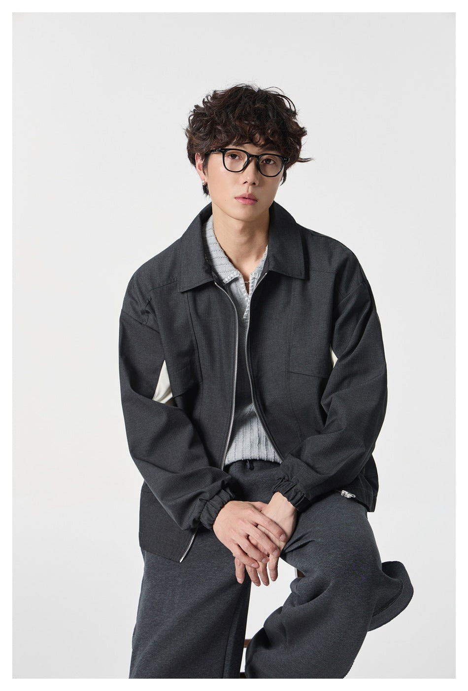 Seated model wearing a charcoal zip-front jacket with a pointed collar and flap chest pockets.