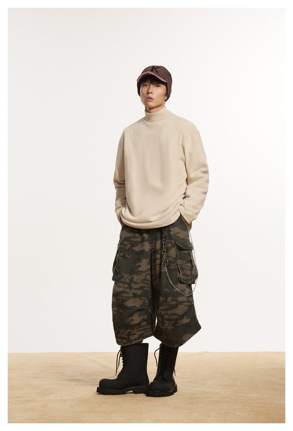 Model wearing beige mock-neck pullover with camouflage cargo shorts, brown cap and black lace-up boots, front view