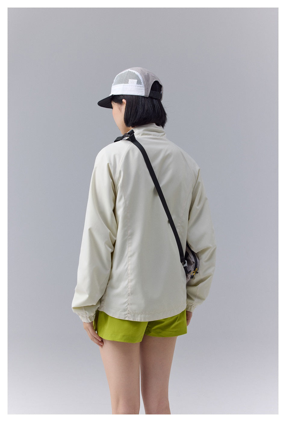 Rear view of a model wearing an ivory outdoor jacket, lime shorts, a mesh cap, and a small crossbody bag.