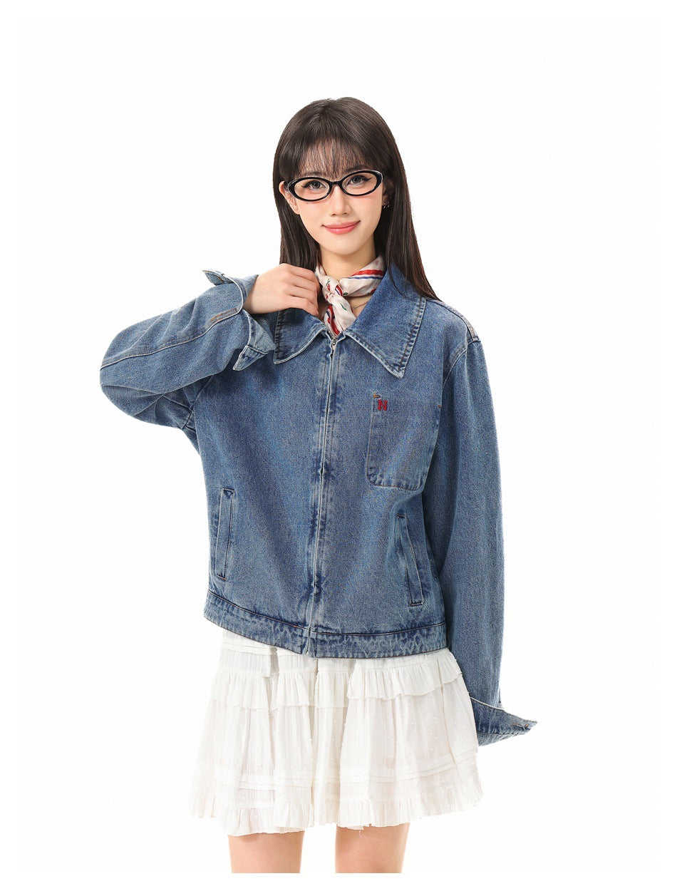 Model wearing a blue denim jacket with a large collar, front pockets and small red embroidery, styled with a white skirt.