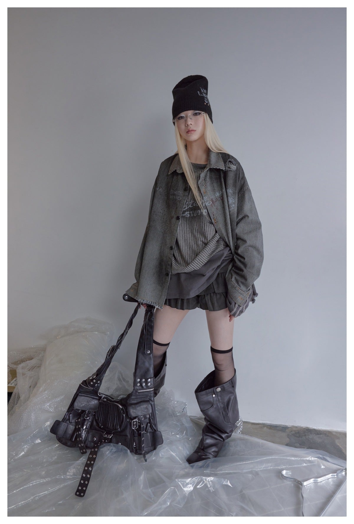 Model wearing an oversized charcoal pinstriped button-up jacket over layered dark tops and shorts, with a beanie and boots.