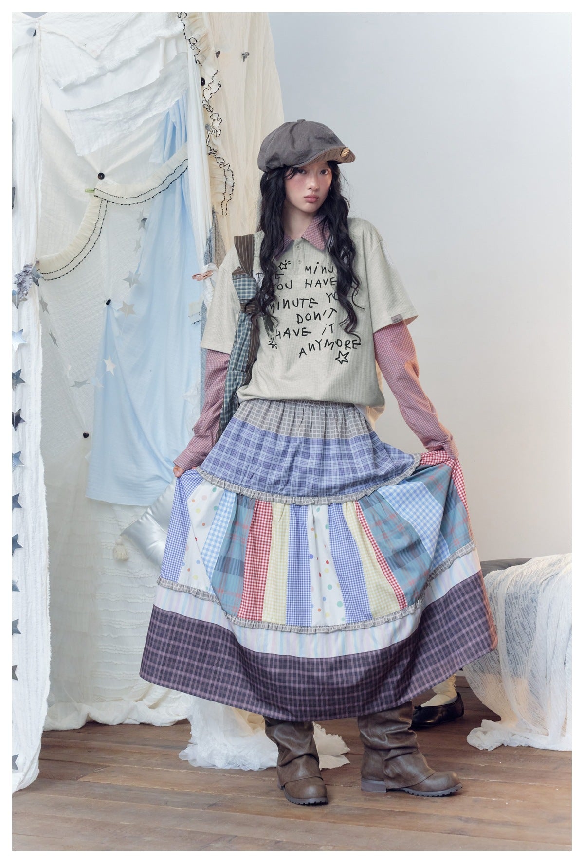 Full-length model wearing a pale gray graphic tee, pink checked shirt, and tiered patchwork plaid skirt.