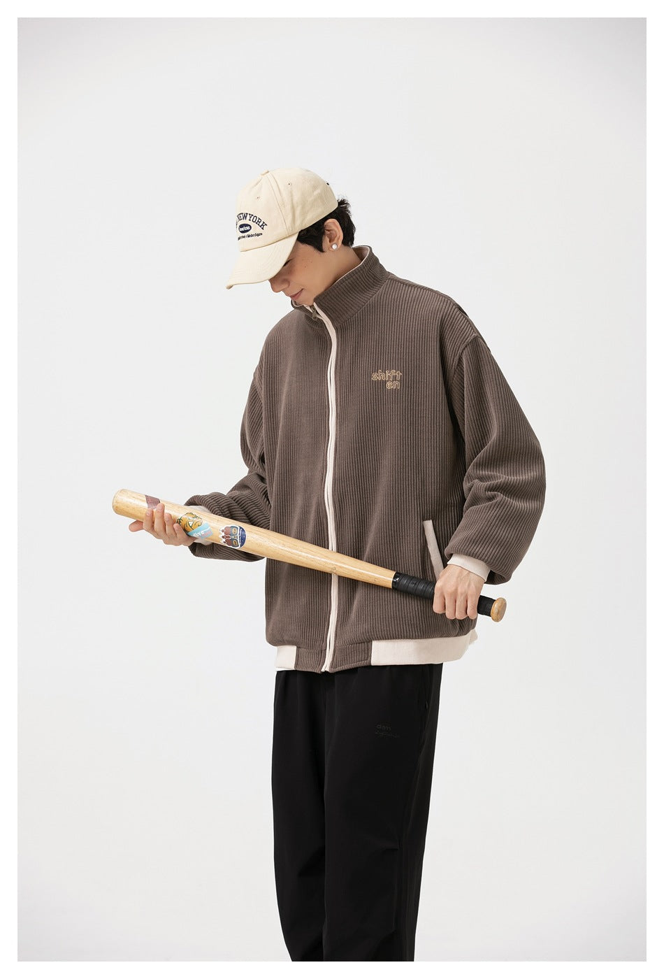 Model wearing the brown ribbed jacket, shown from the side while holding a bat across the front.