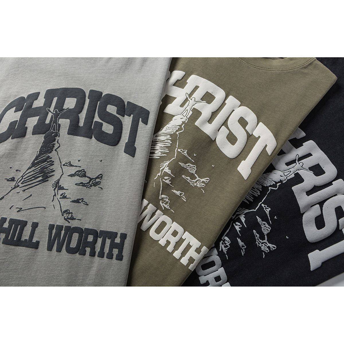 Three overlapping gray, olive, and black T-shirts show a large contrasting religious graphic and text.