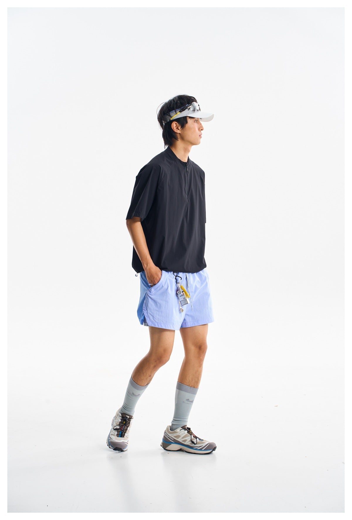 A man wears a black short-sleeve zip-neck top and light blue shorts in a full-body studio photo.