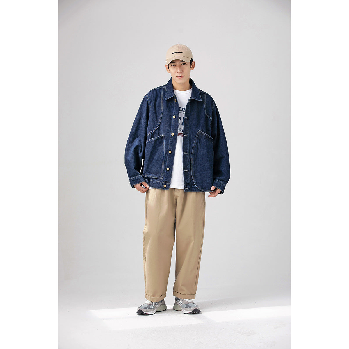 Model wearing a dark indigo denim jacket with large pockets, beige trousers, and a beige cap.