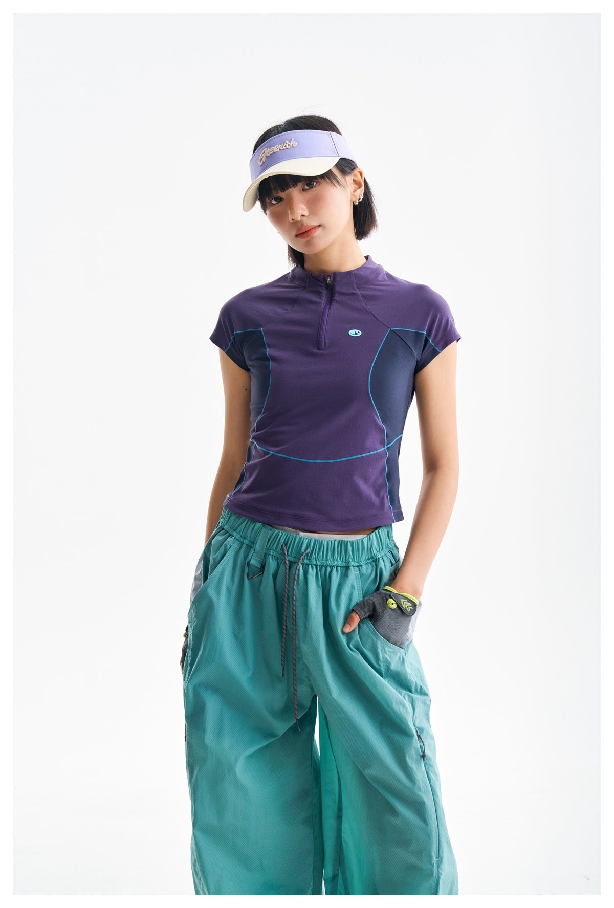 Closer front view of a model in a purple zip-neck top with teal piping and loose teal pants.