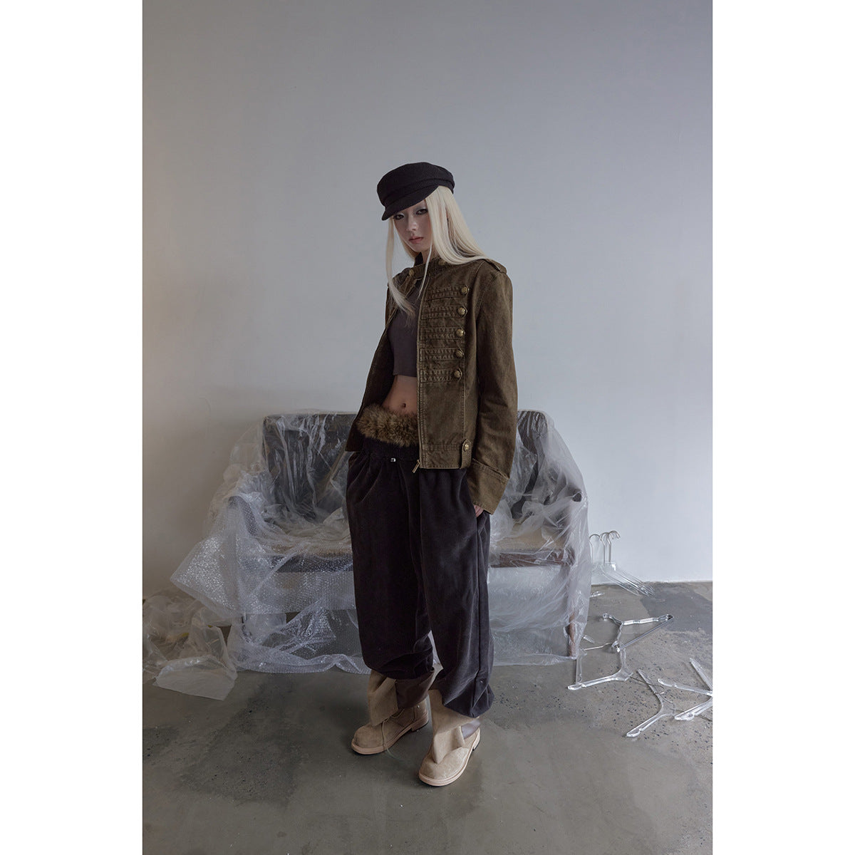 Model wearing a cropped olive-brown military-style jacket with brass buttons, dark loose trousers, and a black cap.
