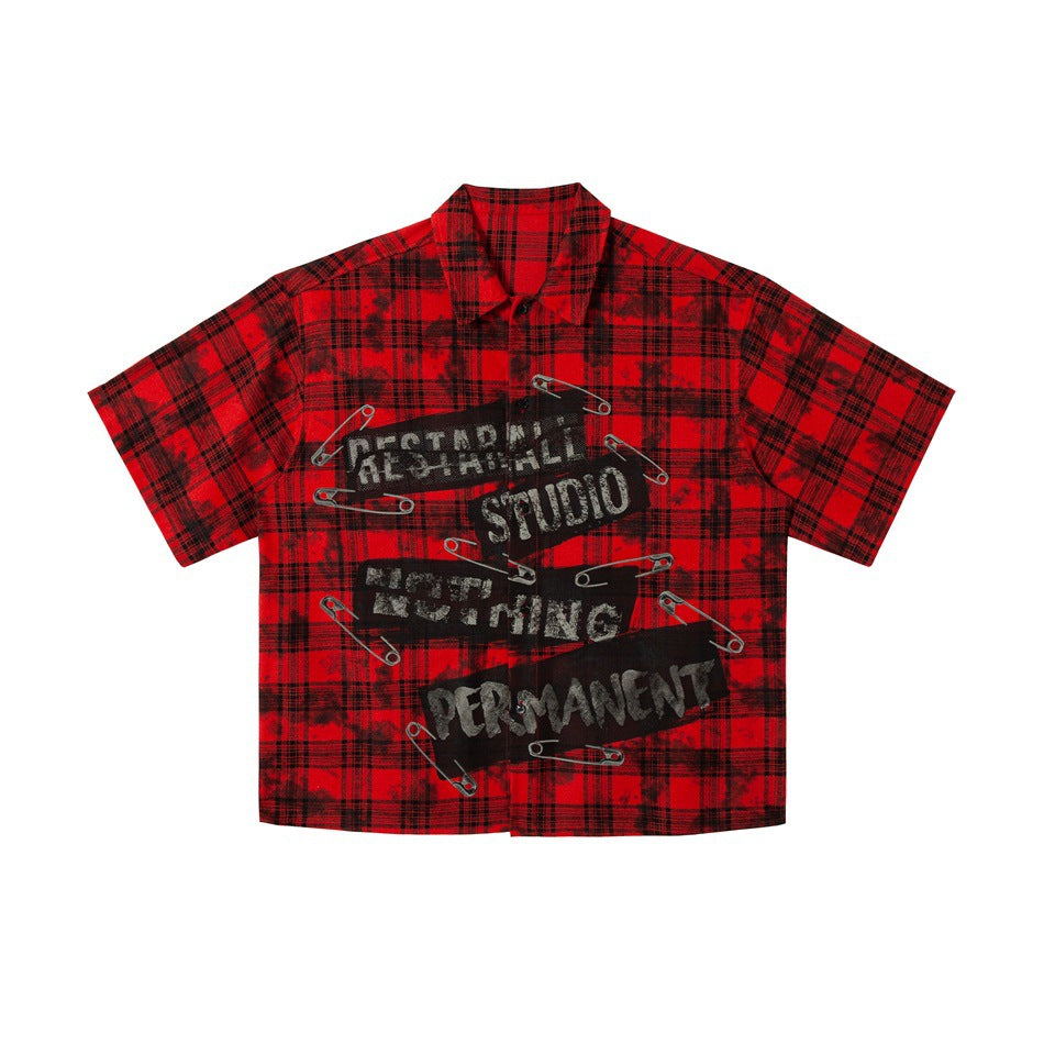 Red and black plaid short-sleeve shirt flat lay, front view with black label patches and safety pin graphics