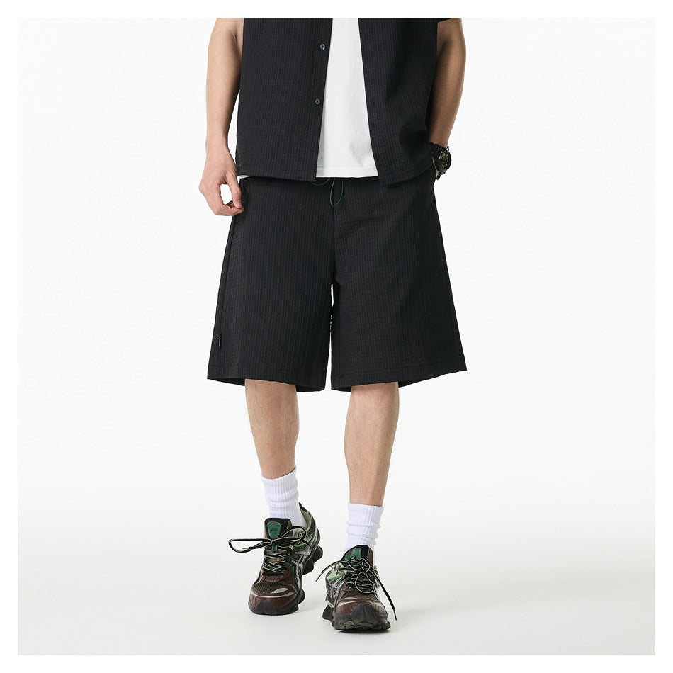 Cropped front view of a model wearing black knee-length shorts with a matching button-front shirt over a white tee.