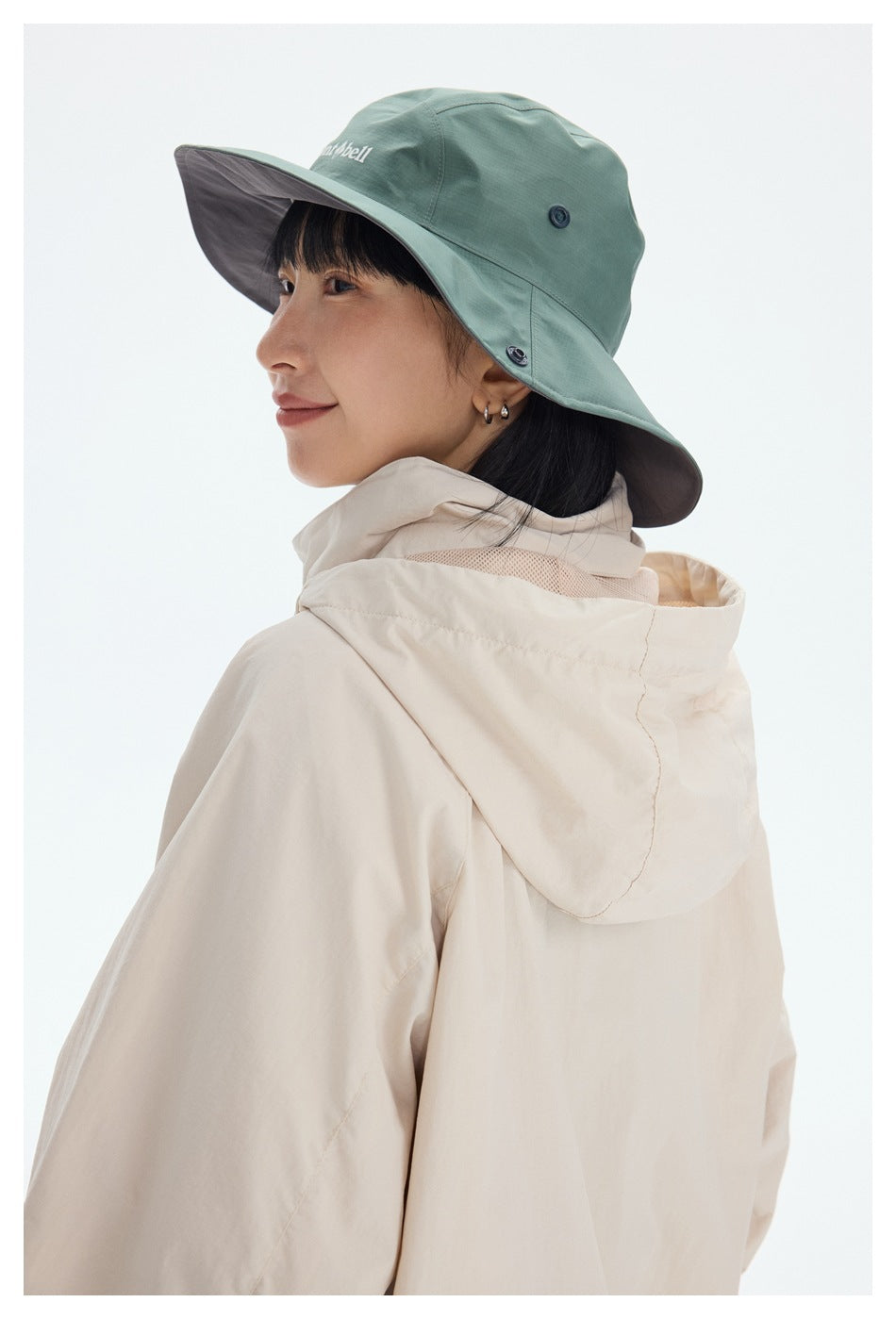 Close-up of a model wearing a cream jacket with a raised hood and a sage-green wide-brim hat.