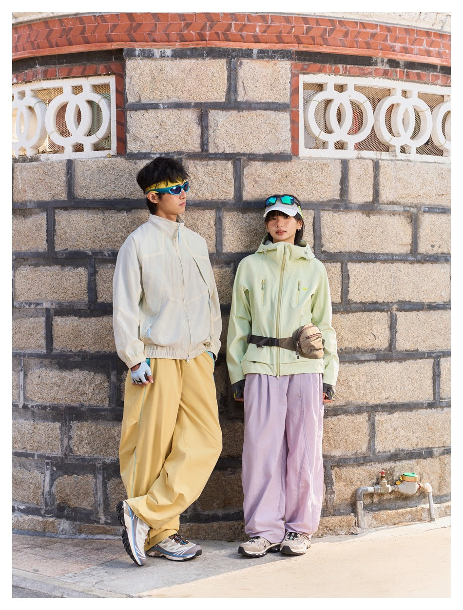 Two models wearing outdoor outfits; the woman wears a pale green hooded jacket with lilac trousers.