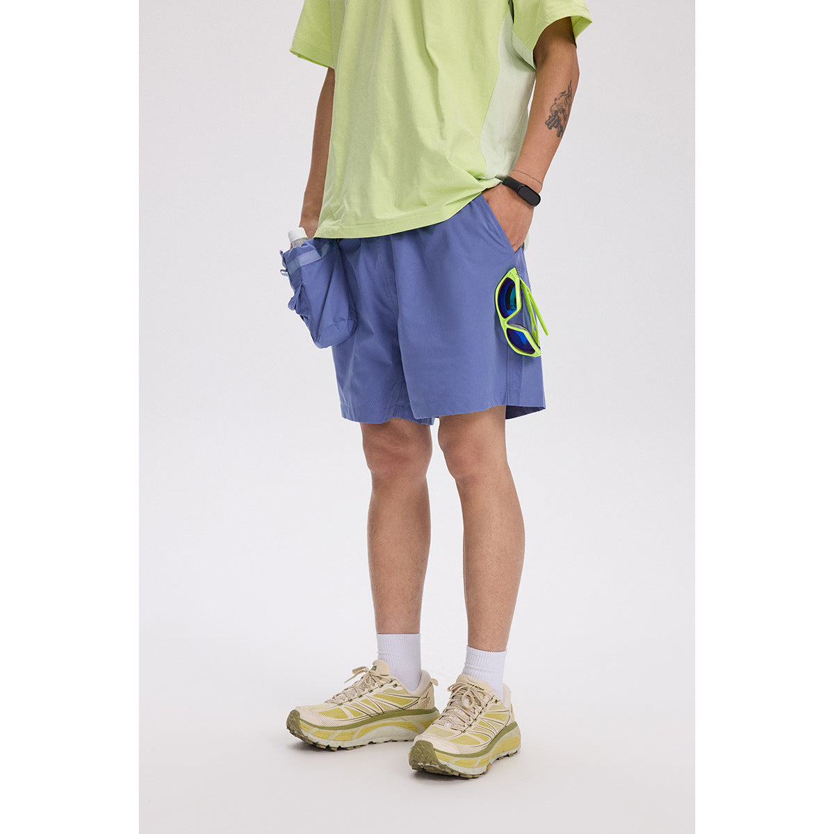 Person wearing blue shorts with a lime-green sunglasses holder clipped at the side, a pale green shirt, and trail shoes.
