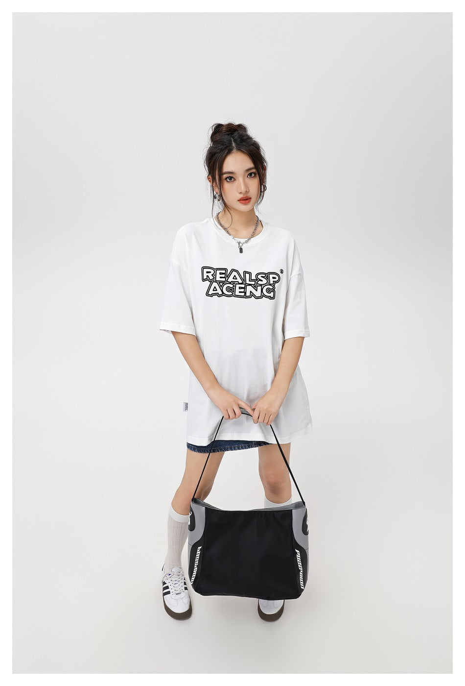Model wearing an oversized white T-shirt with black outlined chest lettering, denim shorts, and a black shoulder bag.