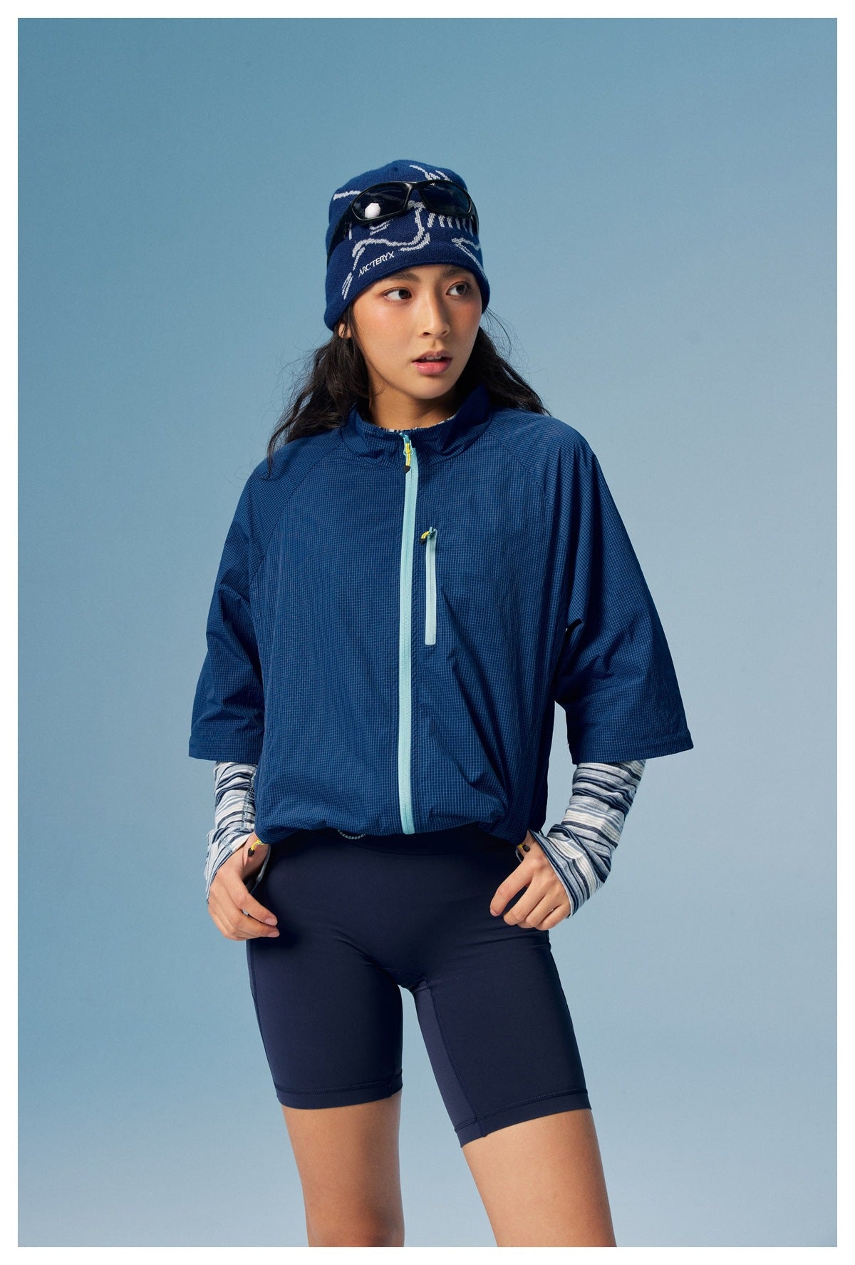 Model wearing a blue short-sleeve zip jacket over striped sleeves, navy cycling shorts, and a patterned blue beanie.