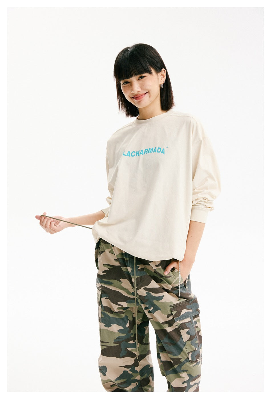 Closer view of a model in an ivory long-sleeve shirt with turquoise chest lettering and camouflage cargo pants.