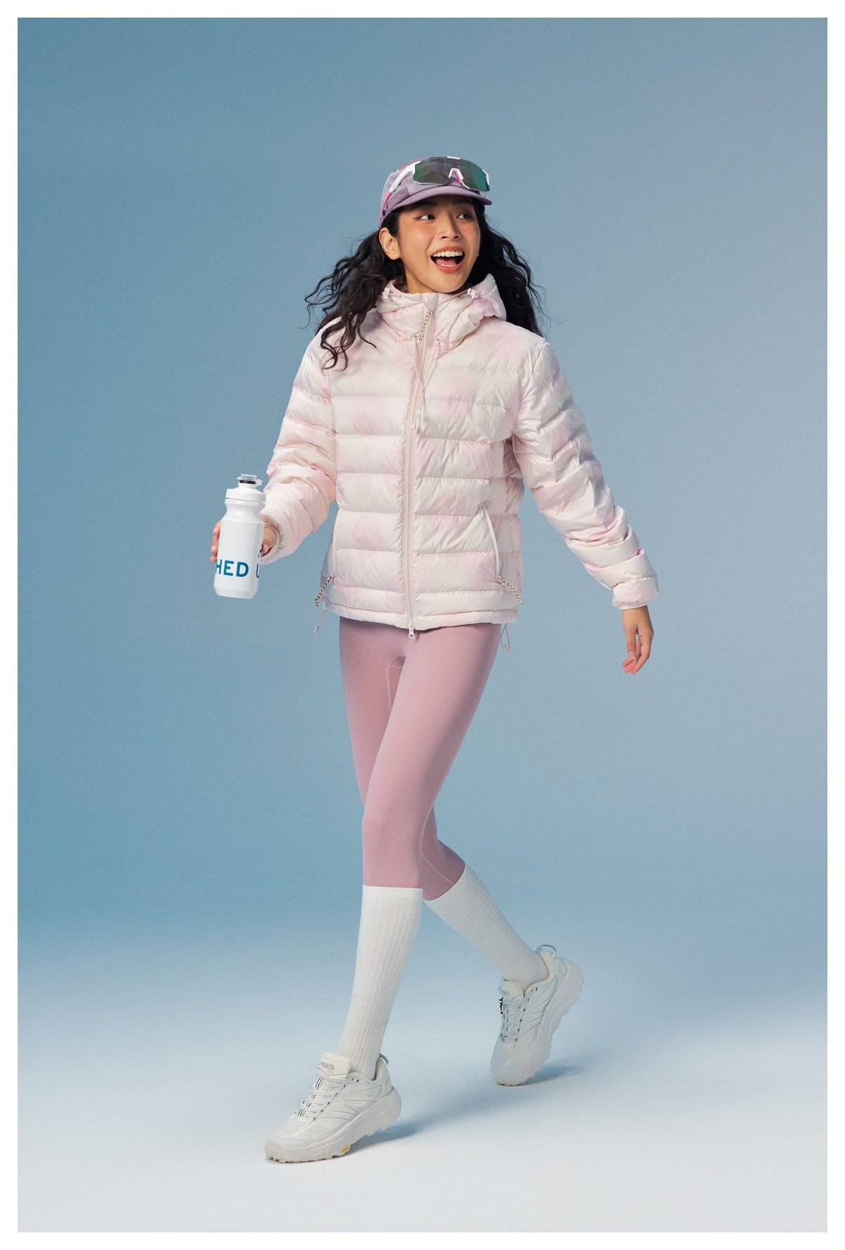Woman walking in the pink hooded quilted puffer jacket with pink leggings and white sneakers, holding a water bottle