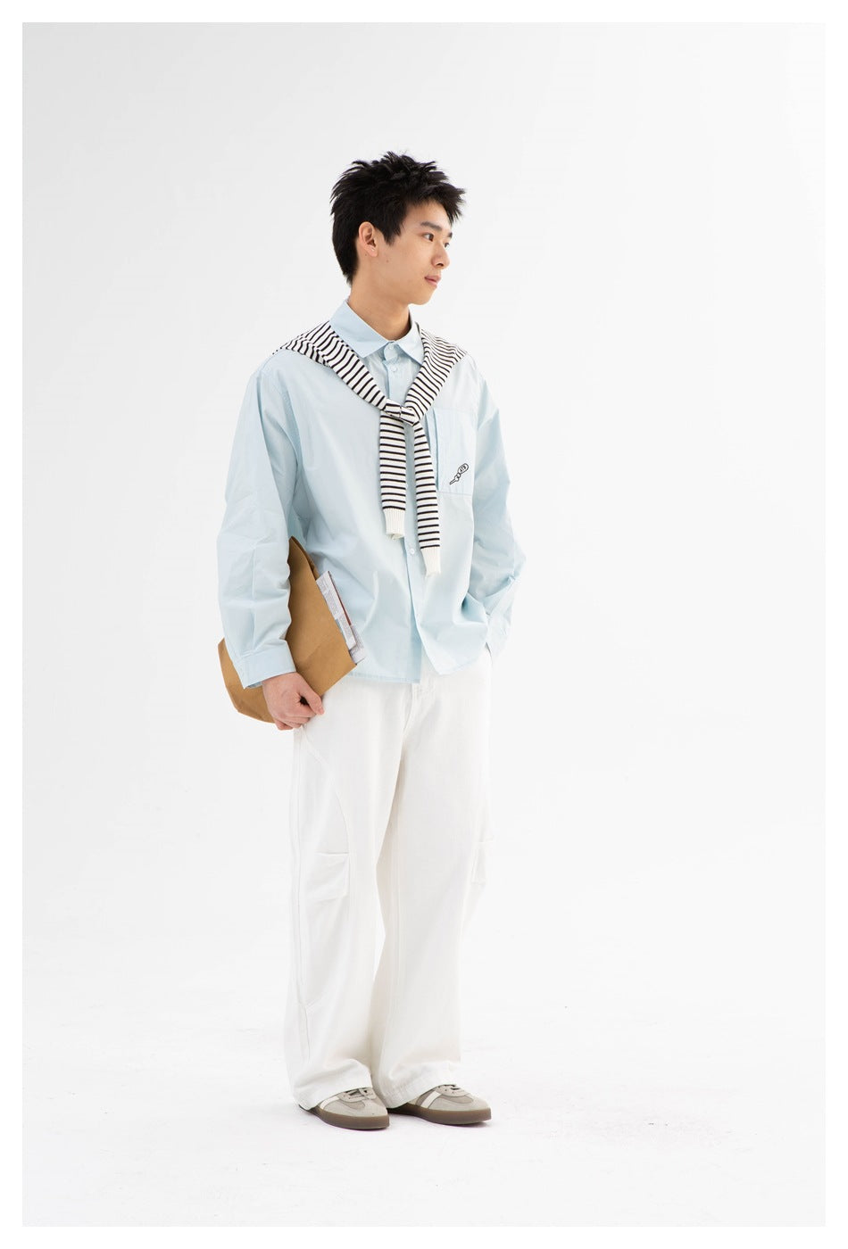 Model in side view wearing a light blue shirt with striped sweater over shoulders, holding a paper bag, with white pants