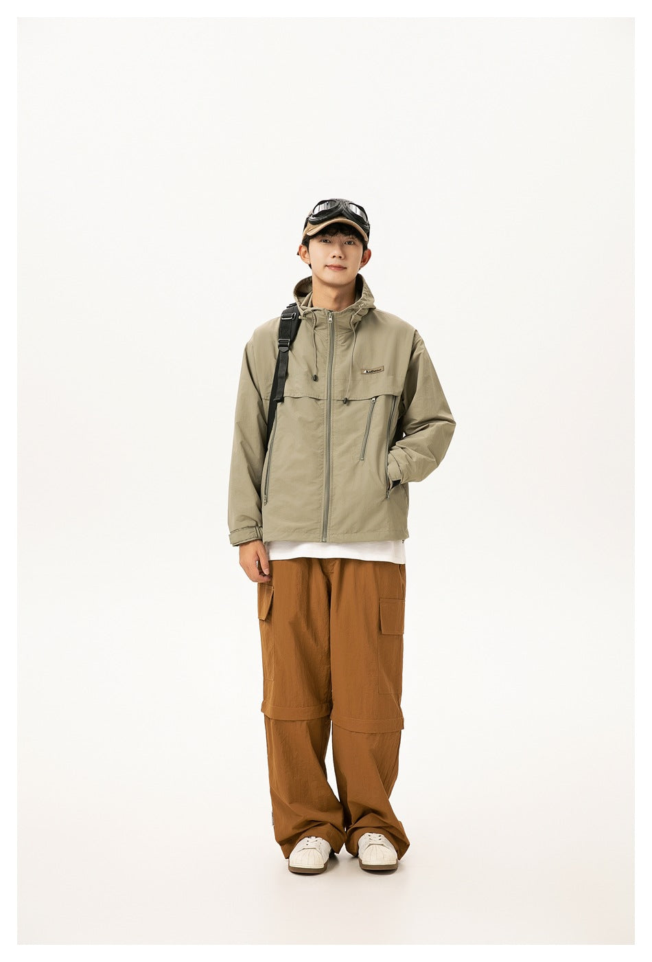 Model wearing light green hooded zip jacket with backpack strap, goggles cap and brown cargo pants