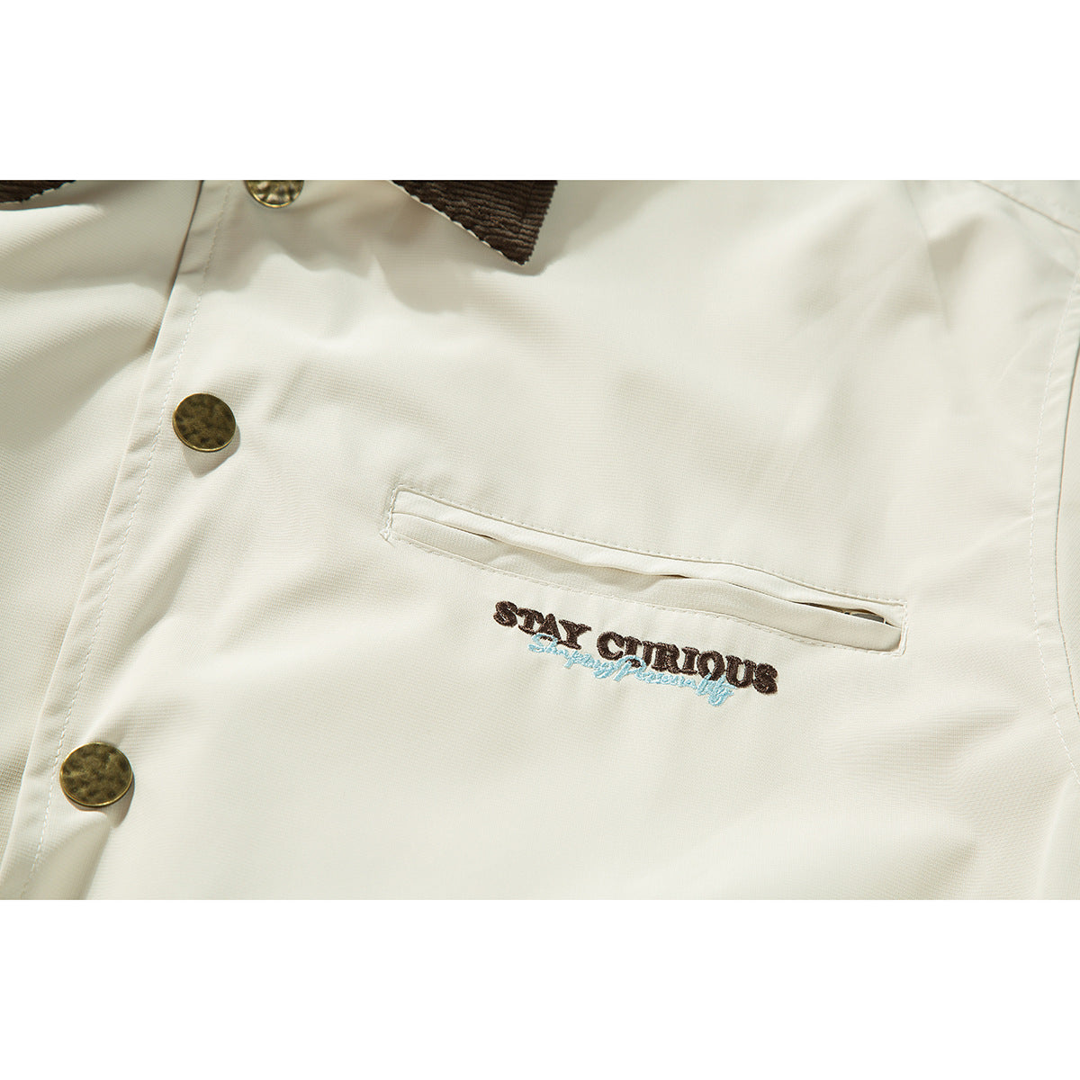 Close-up of an ivory jacket's brass-tone buttons, welt chest pocket, and embroidered lettering.