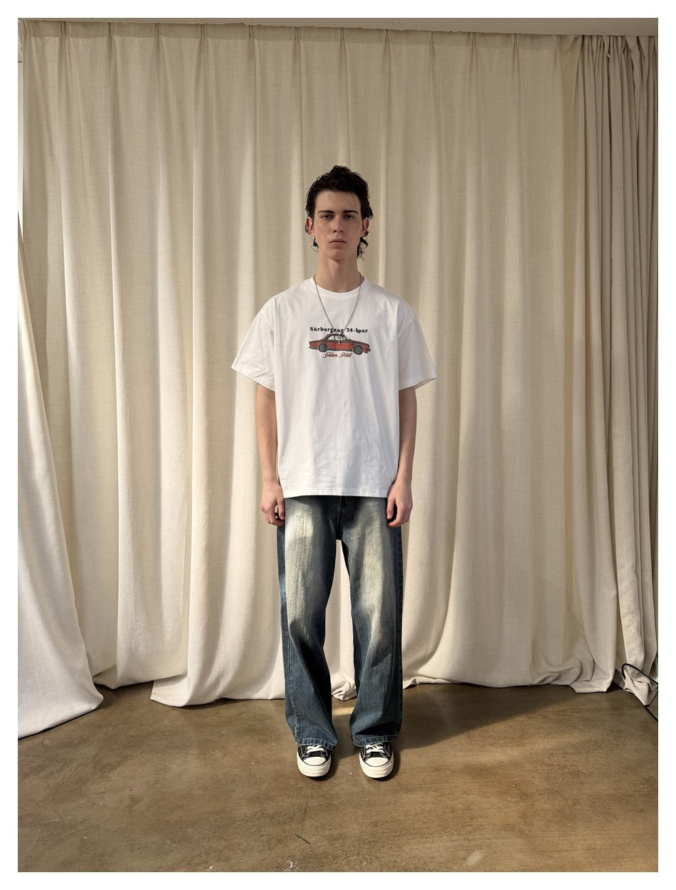 Model wearing a white graphic T-shirt with red car print, wide-leg faded jeans and dark sneakers in front of a cream curtain