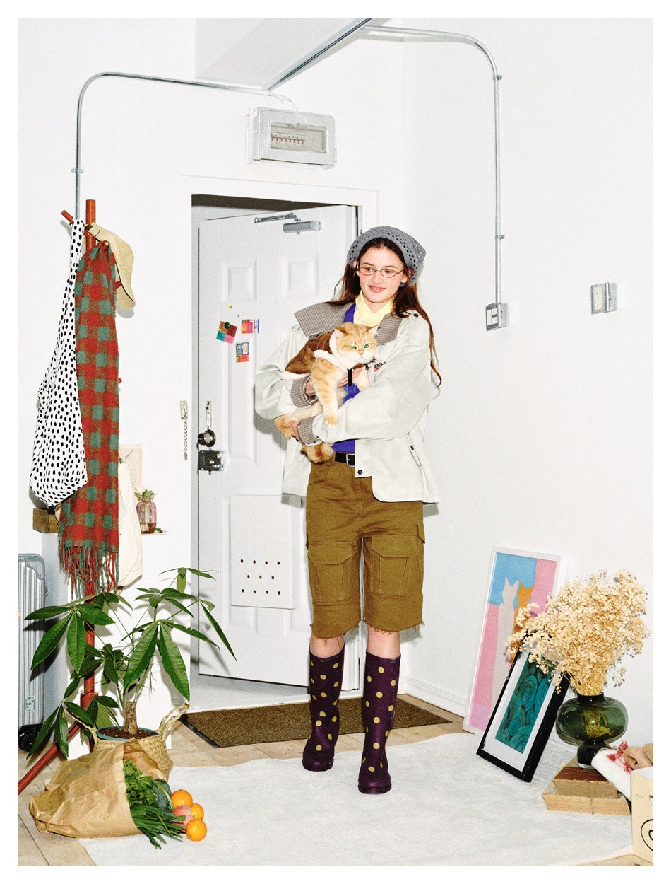 Model holding an orange cat, wearing an ivory jacket with plaid accents, brown cargo shorts, and polka-dot boots.
