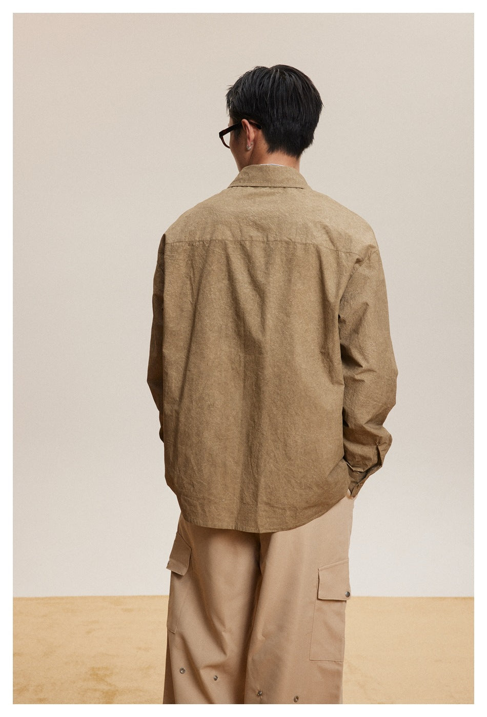 Rear view of a model wearing a loose brown button-up shirt with long sleeves and a pointed collar, paired with beige cargo