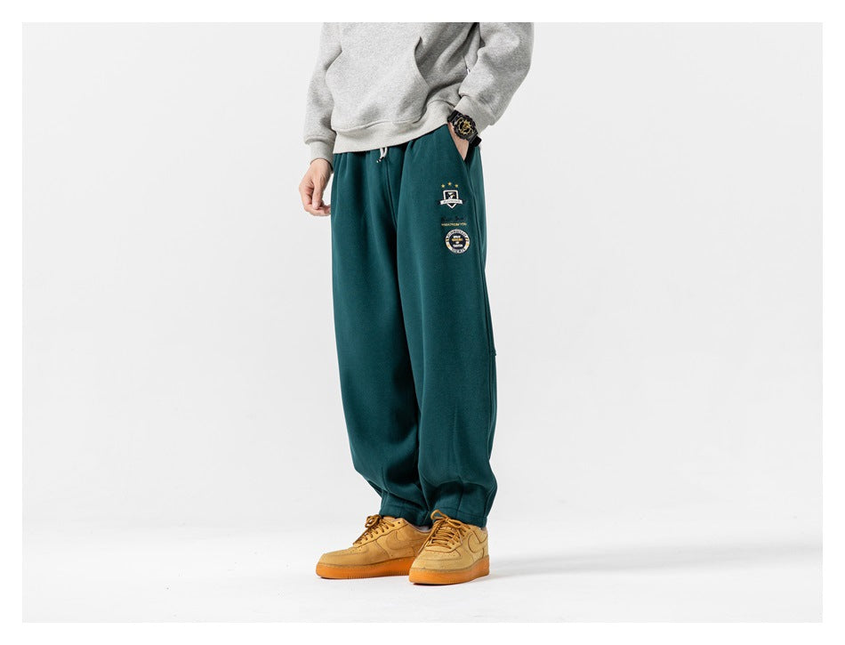 Model wearing dark teal relaxed-fit joggers with an elastic waist, tapered cuffs and small embroidered badges.