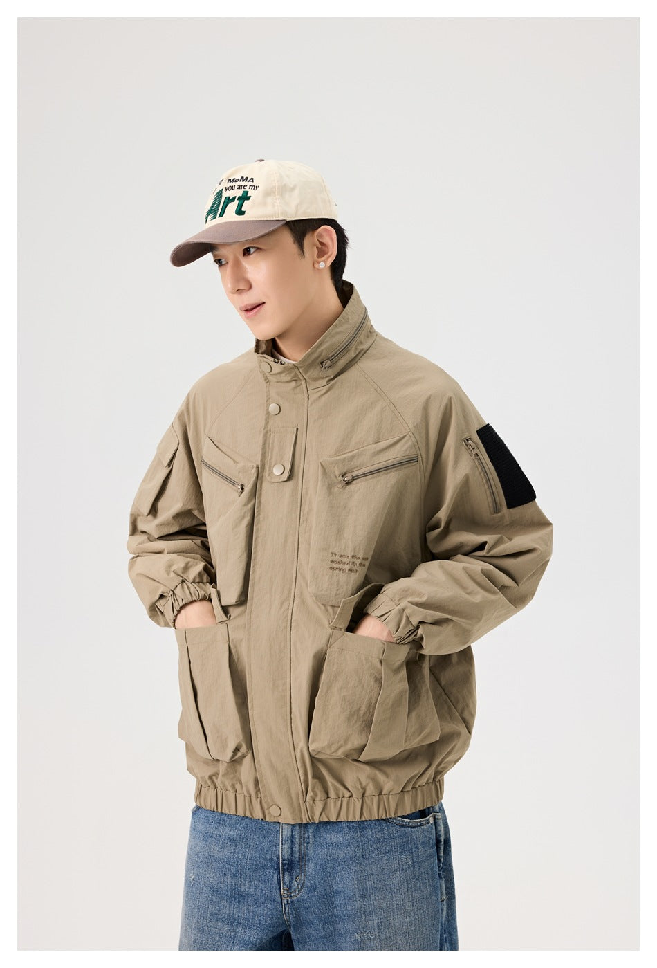 Man wearing a tan utility jacket with a snap-front placket, zip chest pockets, large lower pockets, and a black sleeve patch.