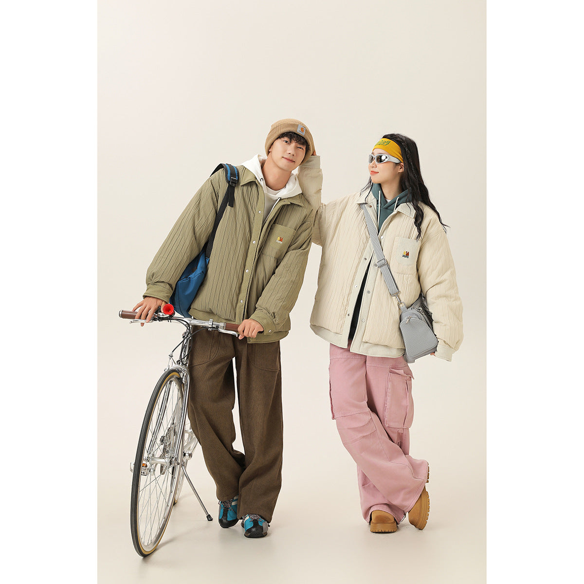 Two models wear quilted snap-front jackets in olive and cream, with a bicycle against a plain studio backdrop.