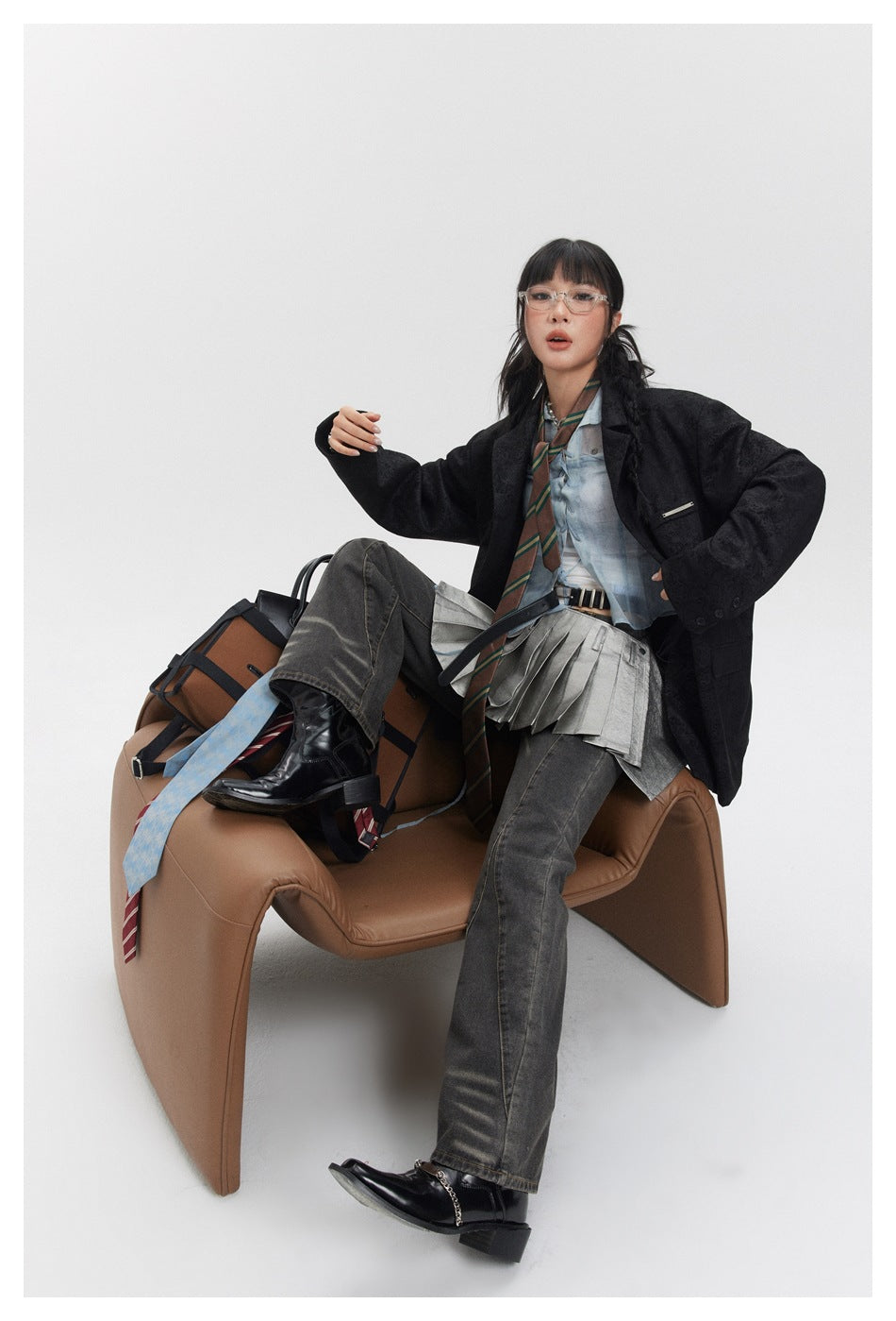 Model seated in a brown chair wearing a black patterned blazer, pale blue shirt, gray pleated layer, and dark jeans.
