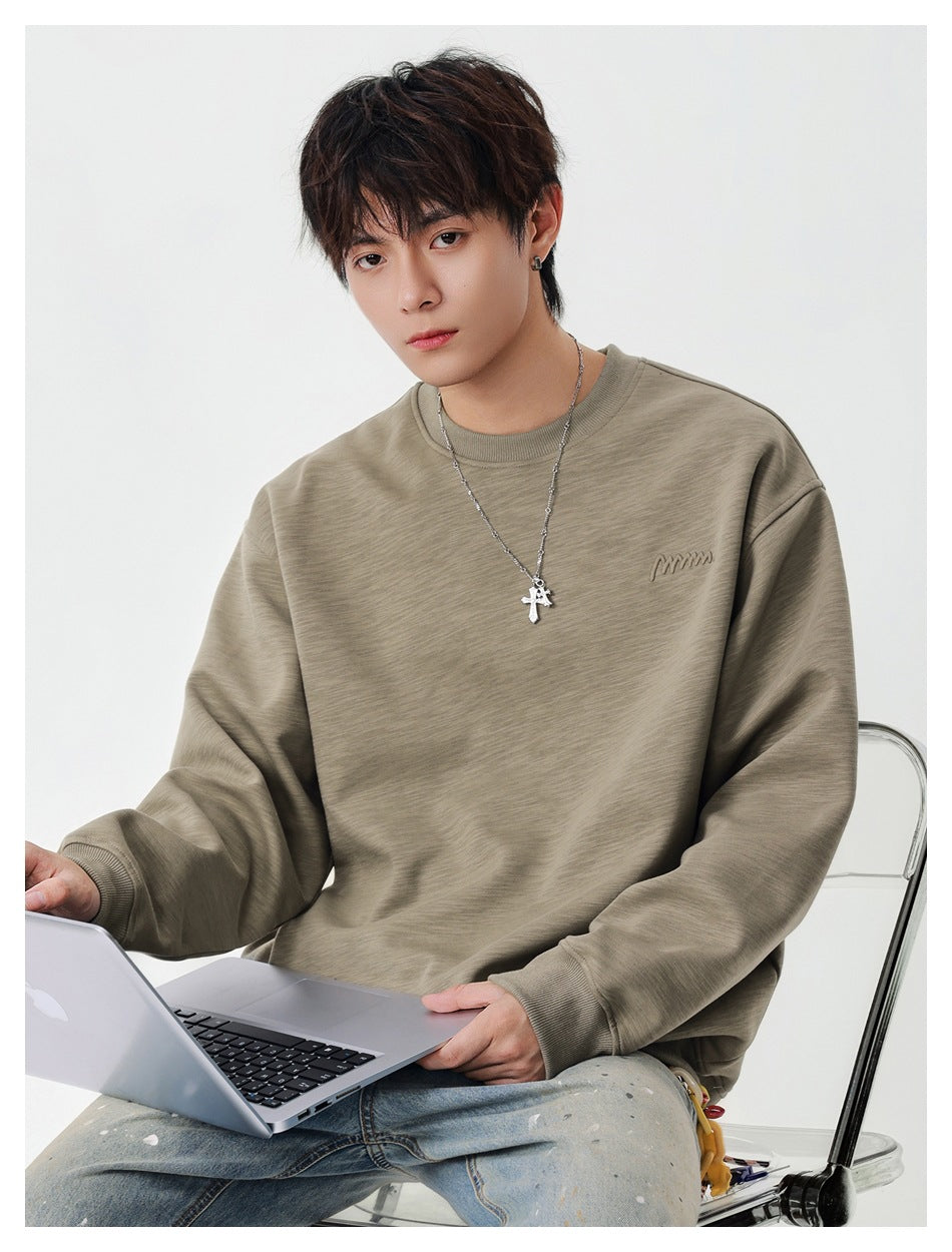 Model seated with a laptop wearing a khaki crew neck sweatshirt with small chest embroidery and cross necklace
