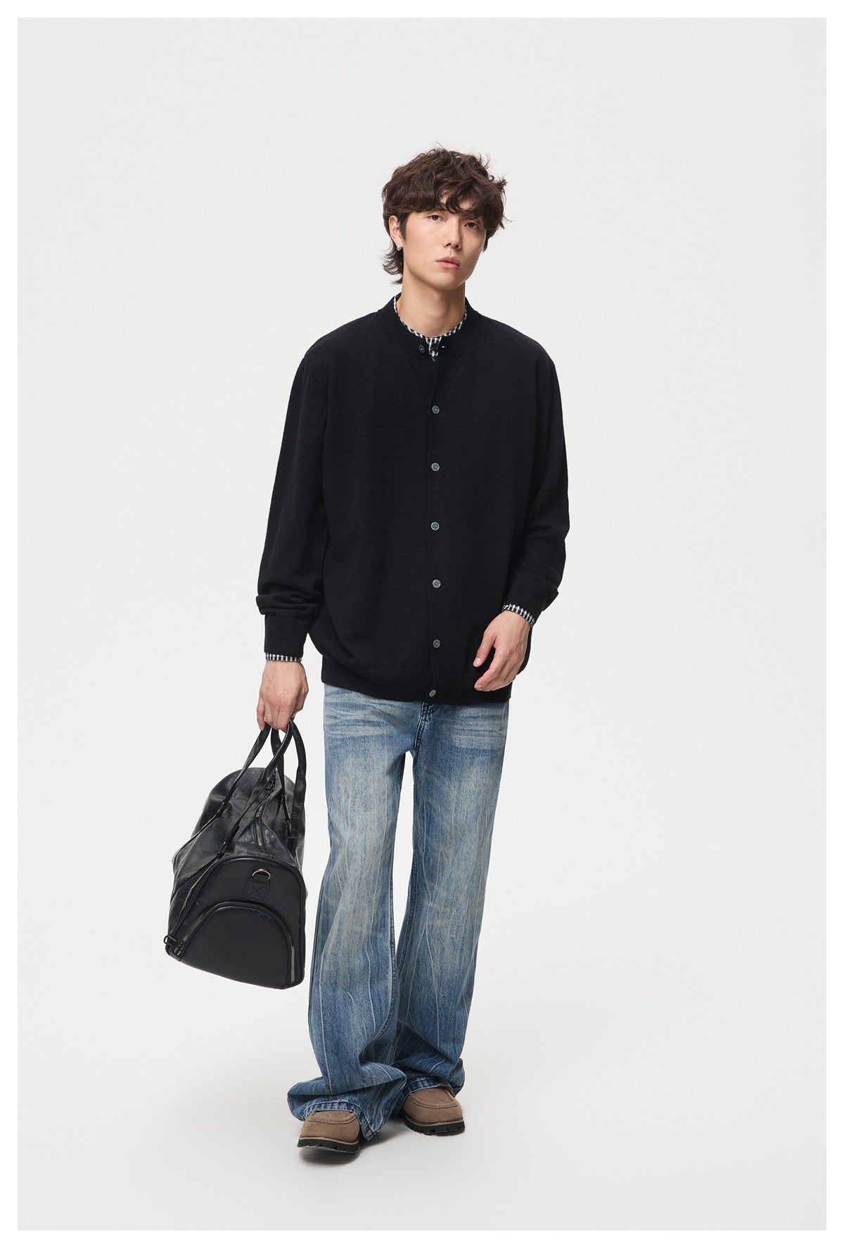 Full-length view of a man in a black button-front cardigan with checked layer, wide-leg jeans and a black duffel bag