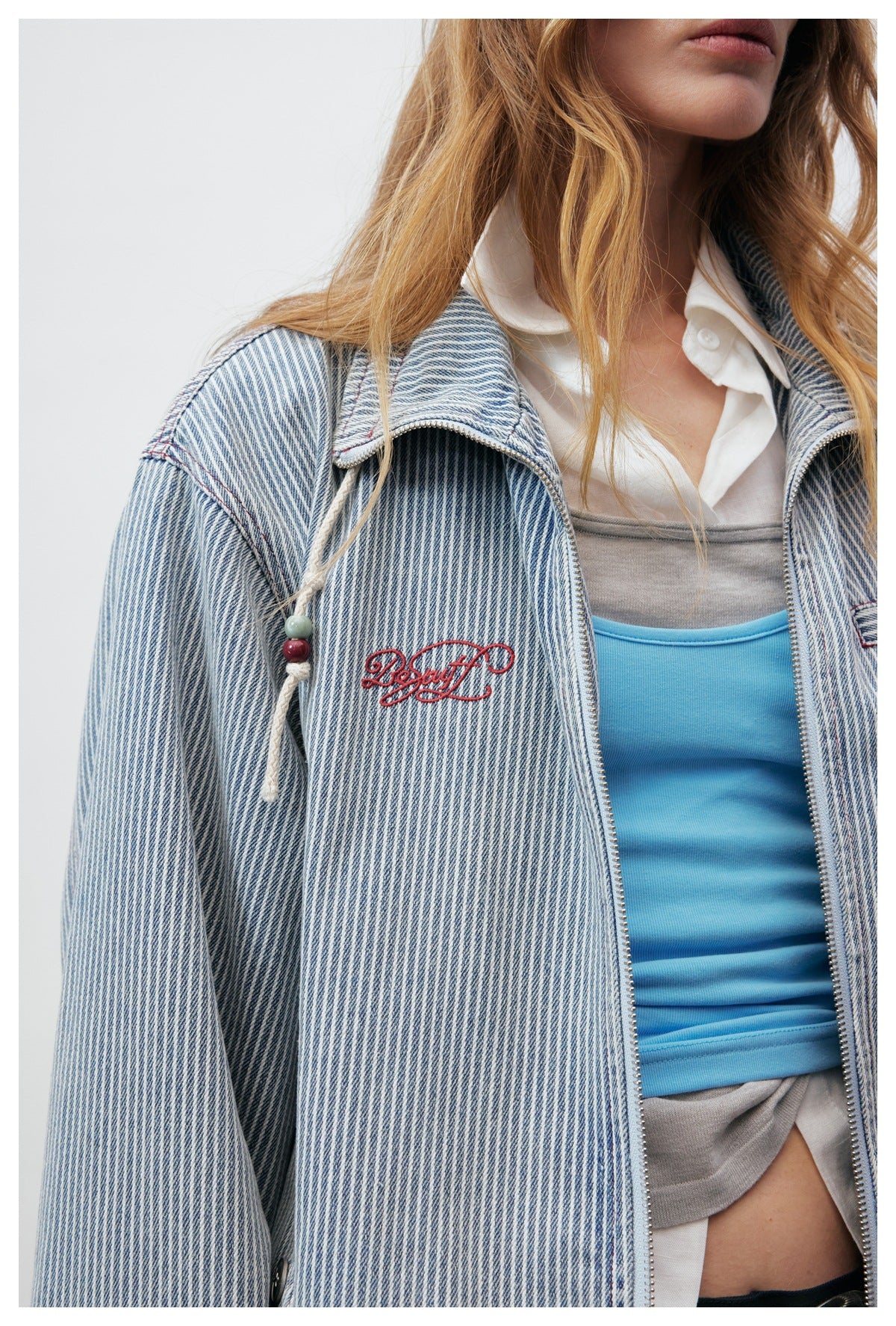 Close view of a model wearing a blue-and-white pinstripe jacket with a zip front, red embroidery and beaded drawcords.
