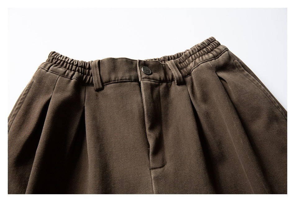 Partial front view of dark brown trousers with a button closure, belt loops, pleats, and elasticated sides.