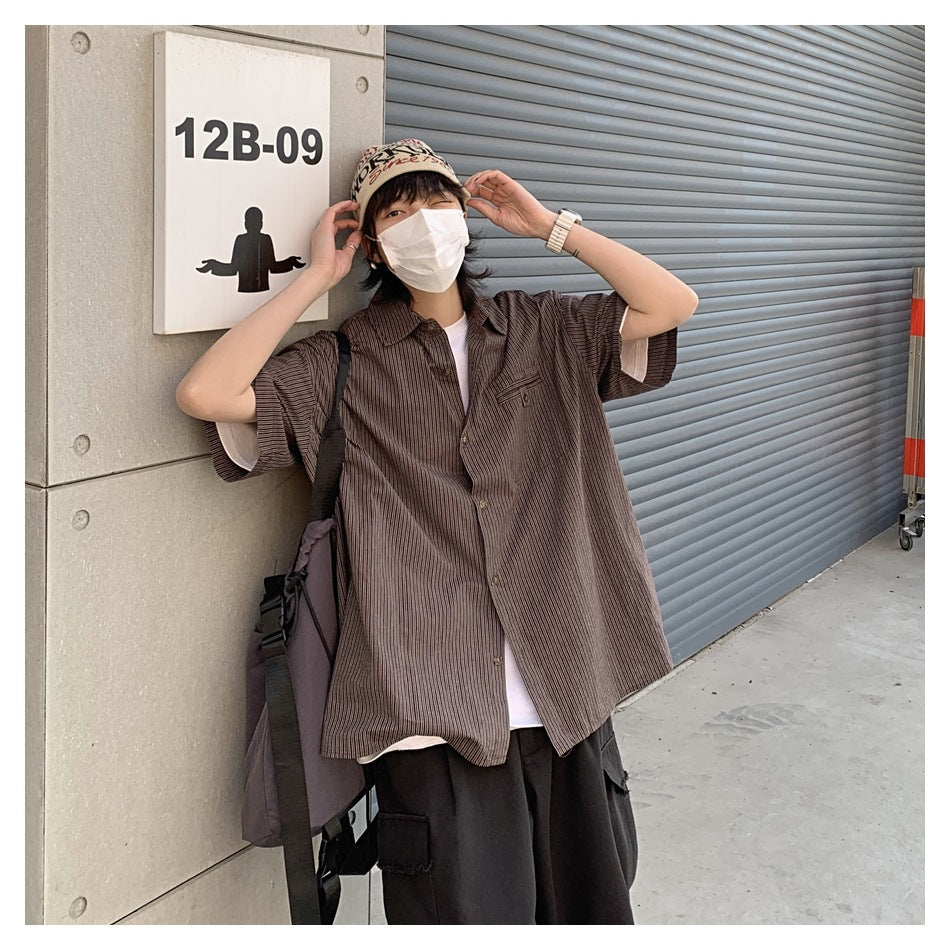Masked model wearing a loose dark brown striped short-sleeve button-up shirt outdoors.