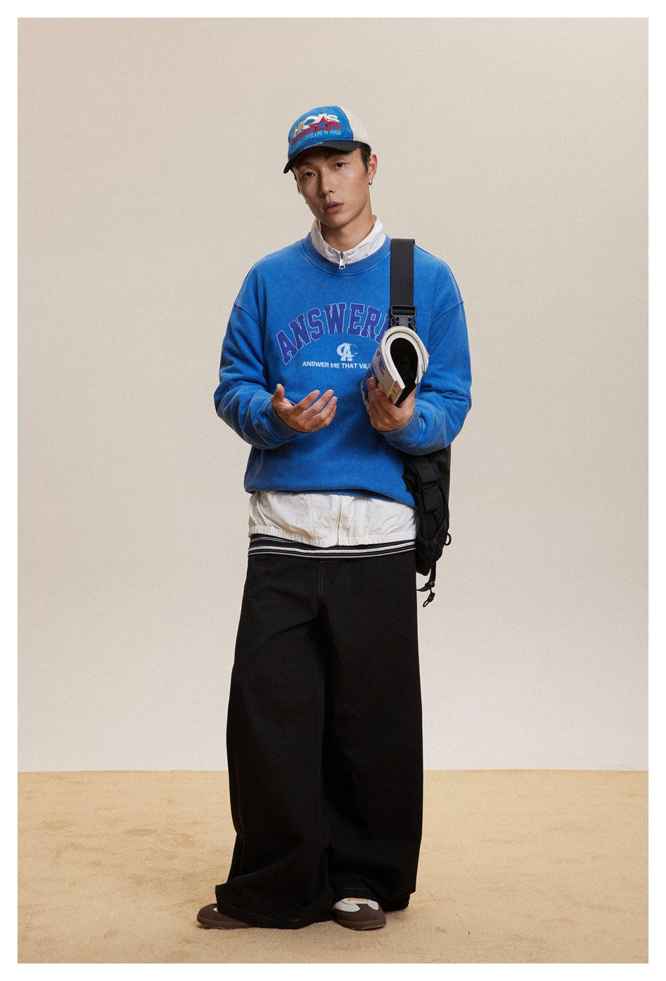 Full-length front view of a model in a bright blue graphic sweatshirt, white jacket, and wide black trousers.