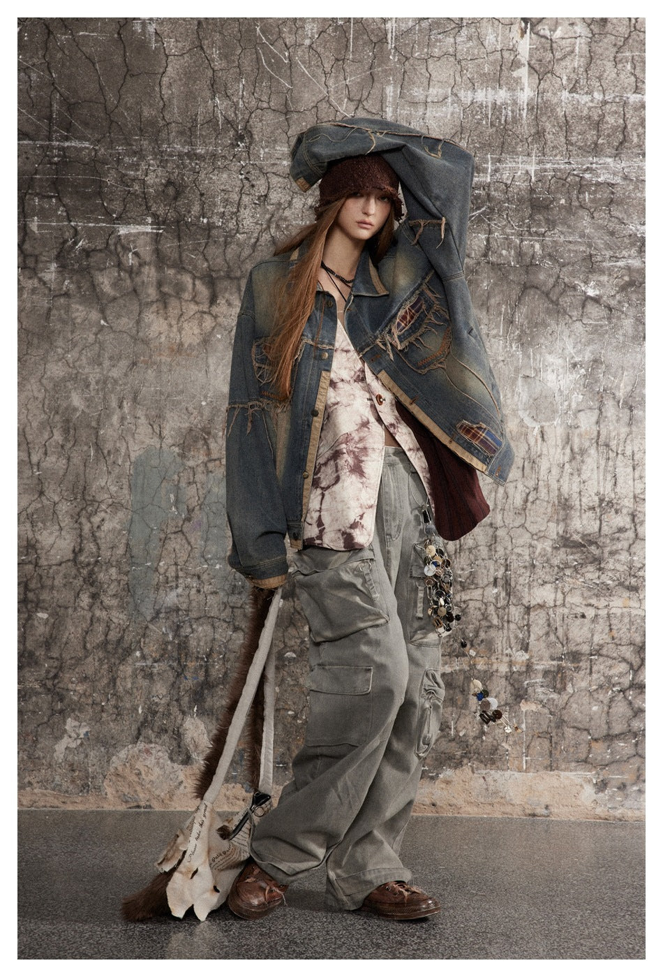 Model poses in an oversized distressed blue denim jacket with plaid patches and loose gray cargo pants.