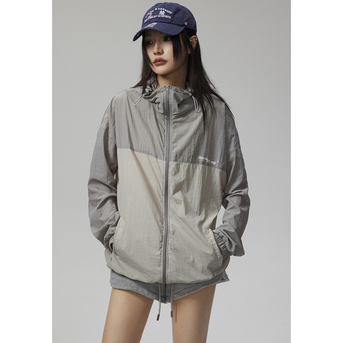 Close view of a model wearing a two-tone light gray hooded zip jacket and navy cap.