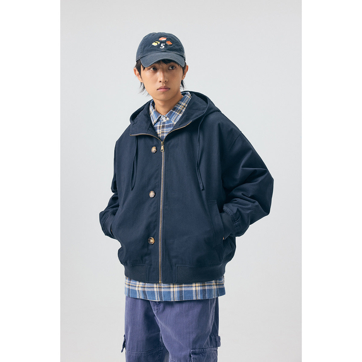 Front view of a model wearing a navy hooded jacket with a zipper, buttons, and pockets over a blue plaid shirt.