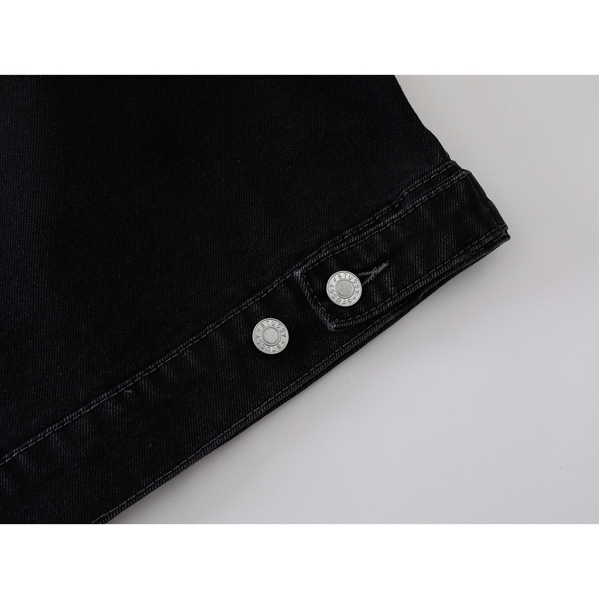 Close-up of a black denim jacket sleeve cuff and hem with silver buttons and contrast stitching.