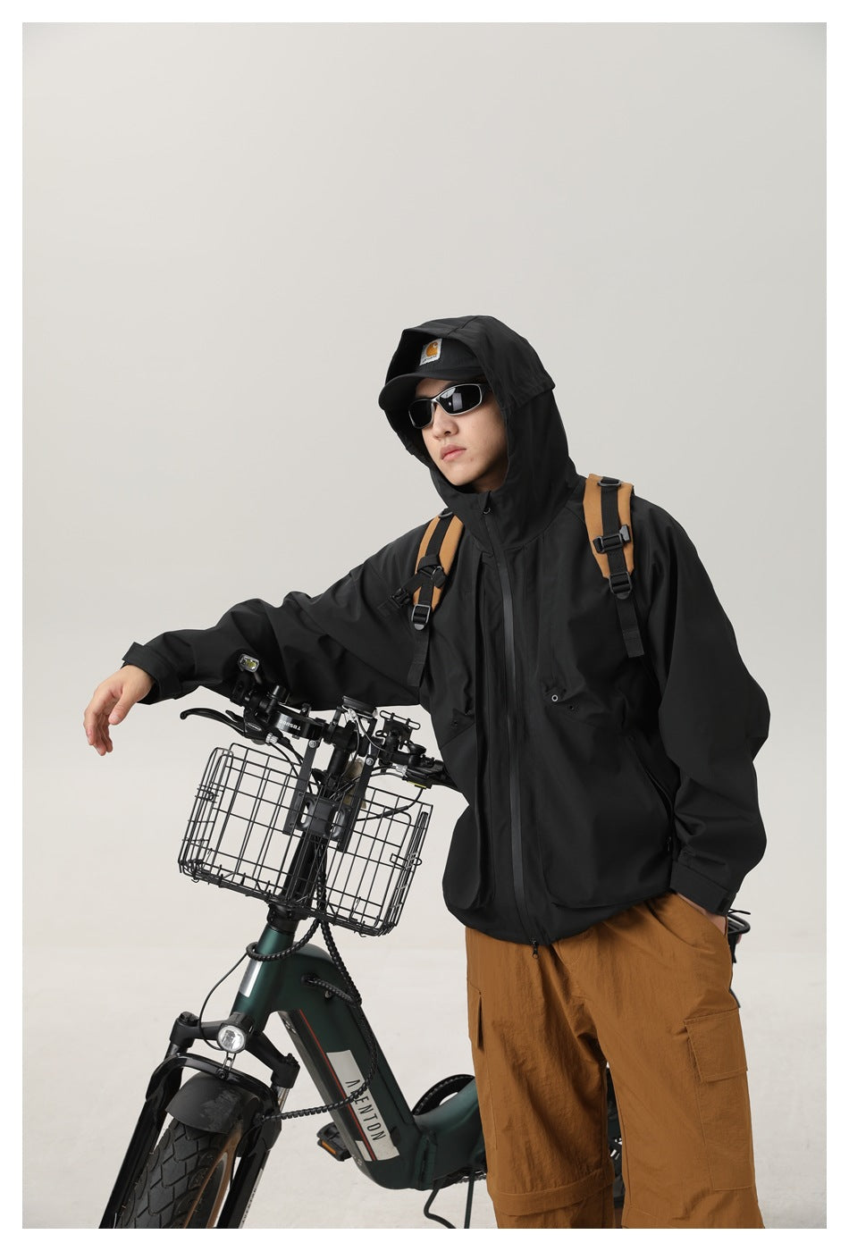 Model wearing a black hooded jacket with a front zipper and flap pockets, beside a bicycle.