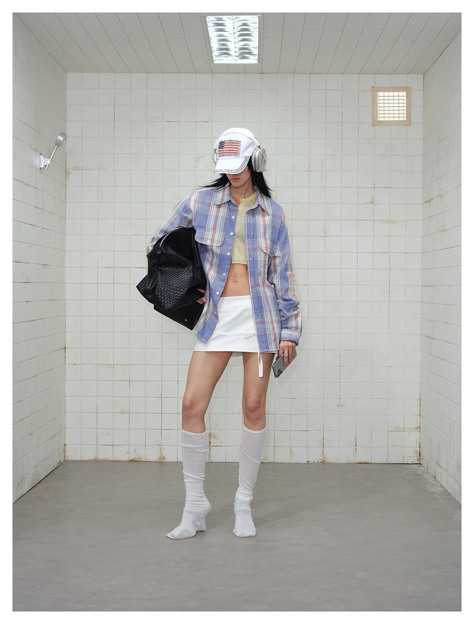Model wearing the plaid shirt open over a cropped top and white mini skirt, holding a black woven bag