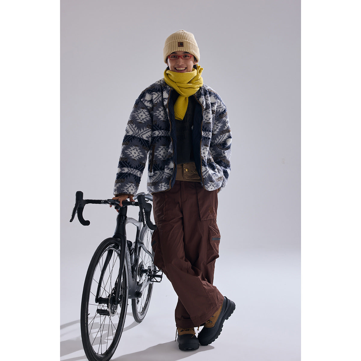 Model wearing an open gray and navy geometric fleece jacket with a yellow scarf and brown cargo pants beside a bicycle.